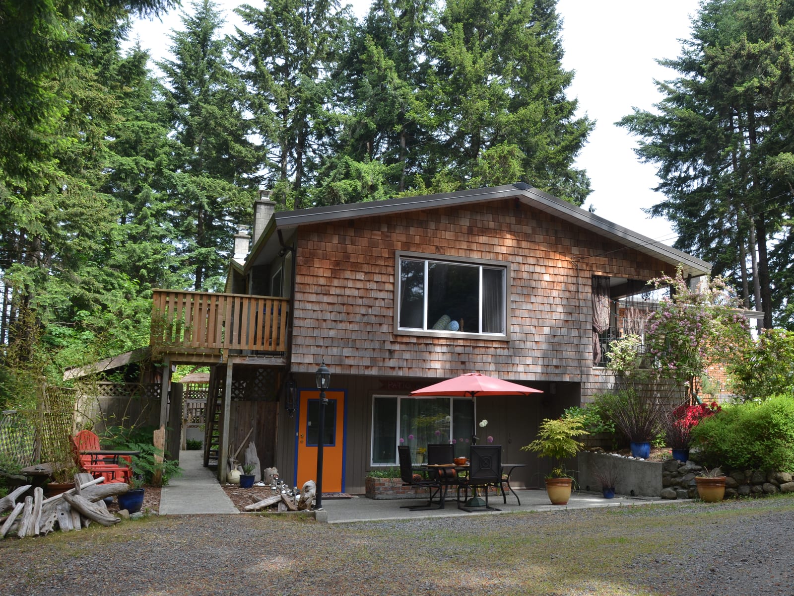 A two-storey cedar-shingled house among tall firs, with a raised deck and a red patio umbrella below it.