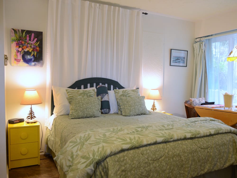A guest room in greens and cream, the bed under a dark carved headboard with a lamp lit on either side.