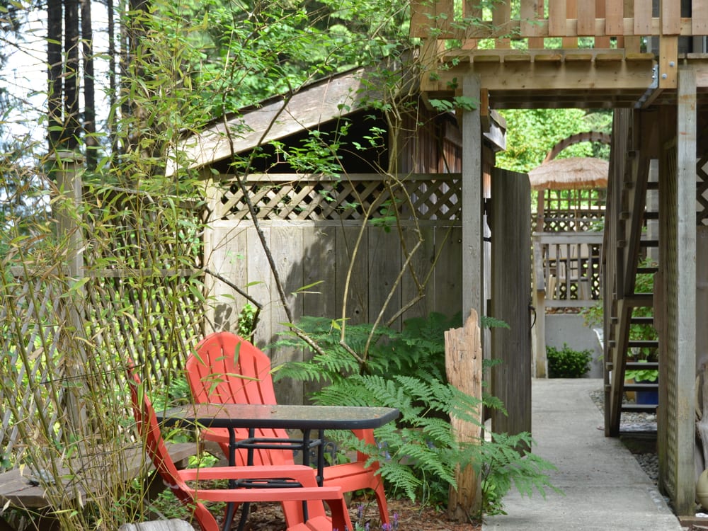 Two red Adirondack chairs under a timber pergola in a corner of the garden, ferns behind them.